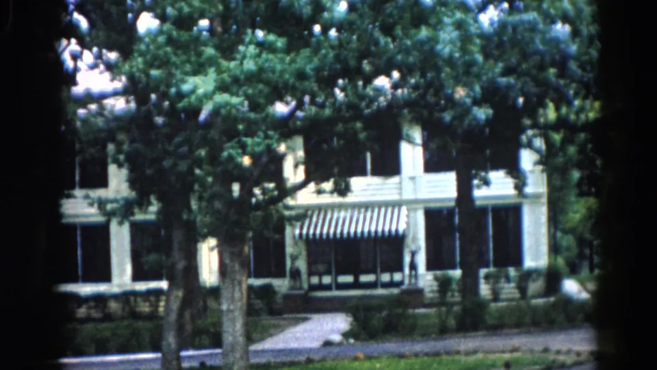 8mm home movie, c. 1962 Illinois — building with awnings seen through tree canopy, with bright film flare or overexposure artifacts in upper foliage.