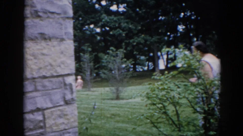 Institutional grounds in Illinois, c. 1962; stone pillar at left frame edge, dense green foliage, and small indistinct figures on the lawn behind.