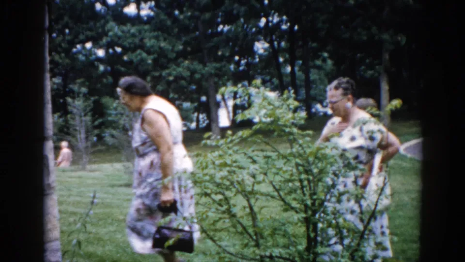 Two women in 1960s patterned summer dresses walking on institutional grounds in Illinois, c. 1962; one carries a dark handbag; a third figure is visible in the background.