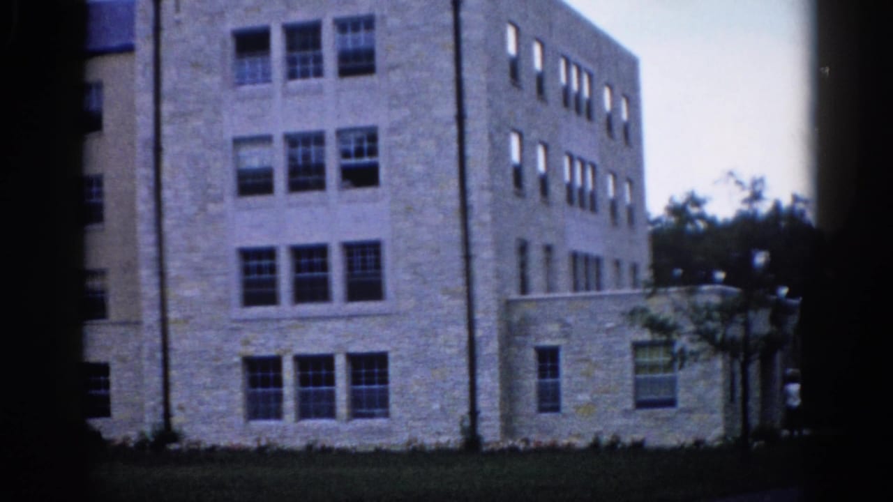 Four-story institutional building on an Illinois campus, circa 1962, shot on 8mm through foreground tree trunks; flat roof and grouped rectangular windows visible.