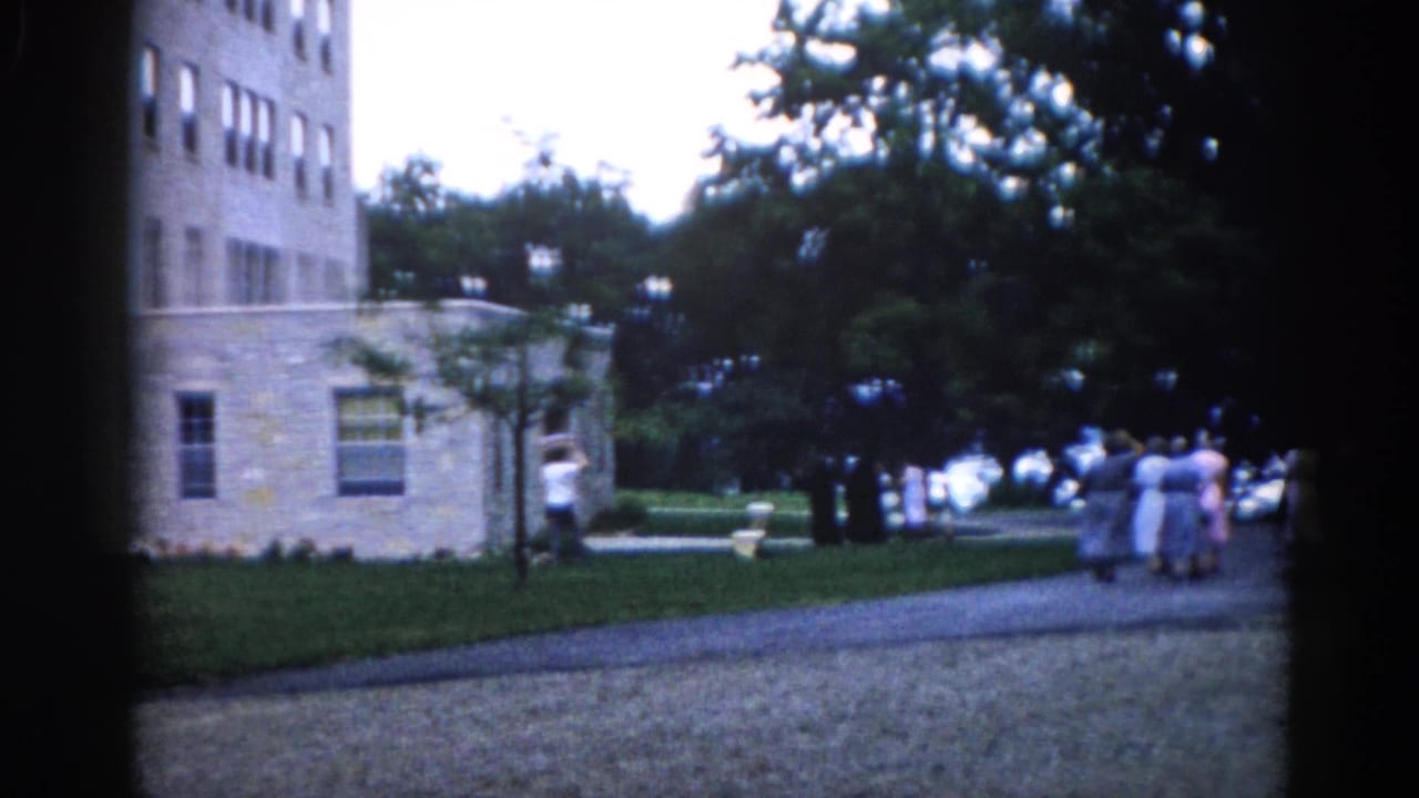 Group of women in mid-century dresses walking away along a gravel path beside a large light stone multi-story institutional building.