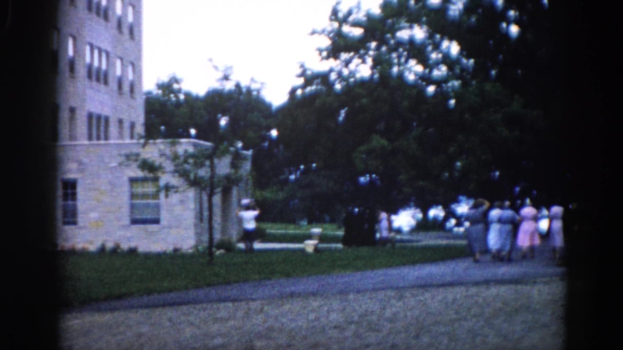 Women continuing along the gravel path beside the stone building, with a lone figure standing to the left near the lawn edge.