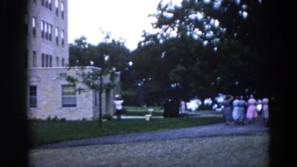Women in early-1960s dress on a gravel path beside a stone multi-story building in Illinois, with a solitary figure to the left; 8mm home movie circa 1962.