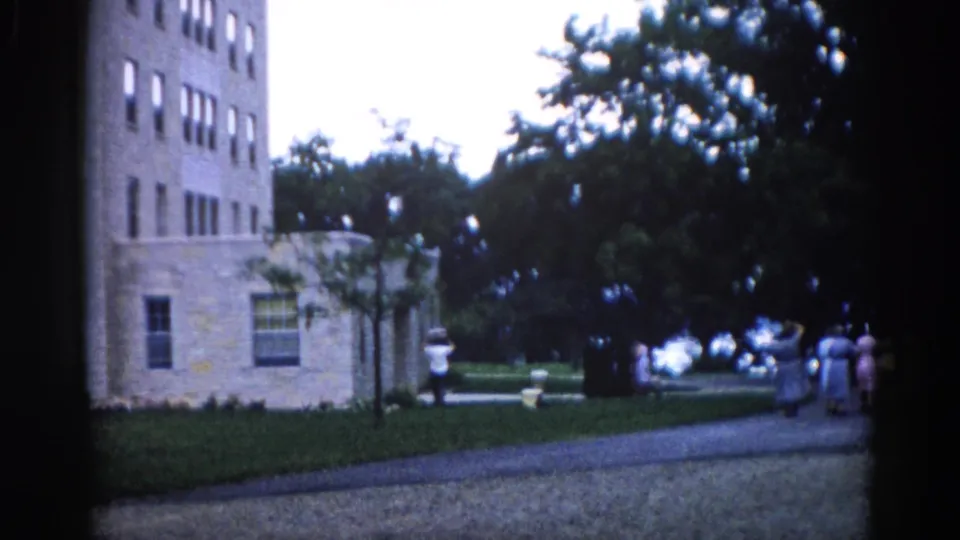 8mm home movie frame circa 1962 Illinois: women receding along a path beside the tall stone building, with prominent lens-flare artifacts and film grain.