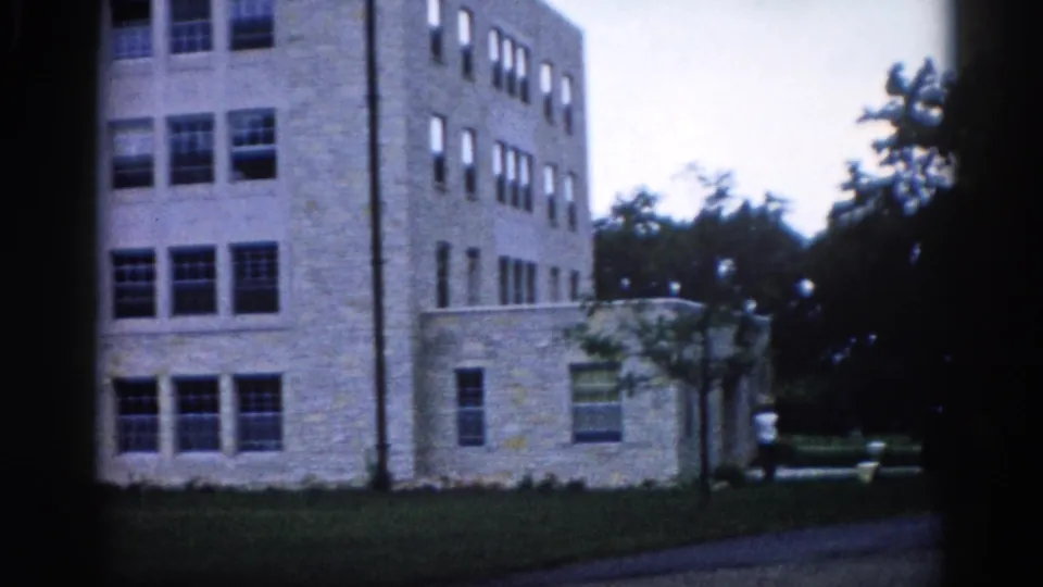Lower facade of a multi-story light stone institutional building in Illinois, circa 1962, showing regular grid-pattern windows, a lamp post, and a lone figure at the base — 8mm home movie.