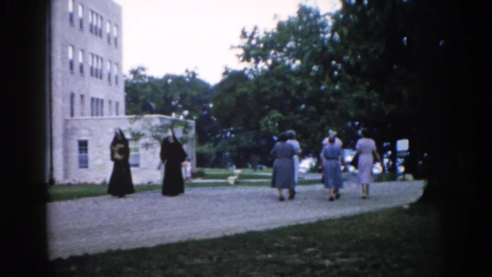 8mm film, c.1962, Illinois — women in religious habits and laypeople on a gravel path beside a multi-story stone convent building amid green summer grounds.