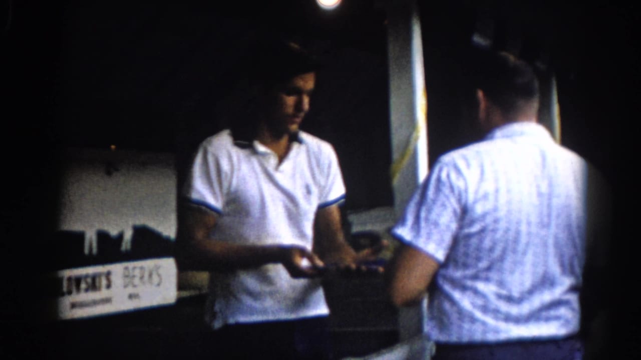 Vendor in white polo faces customer in striped shirt at dim-lit wooden booth; partial business sign at left, yellow banner visible upper right.