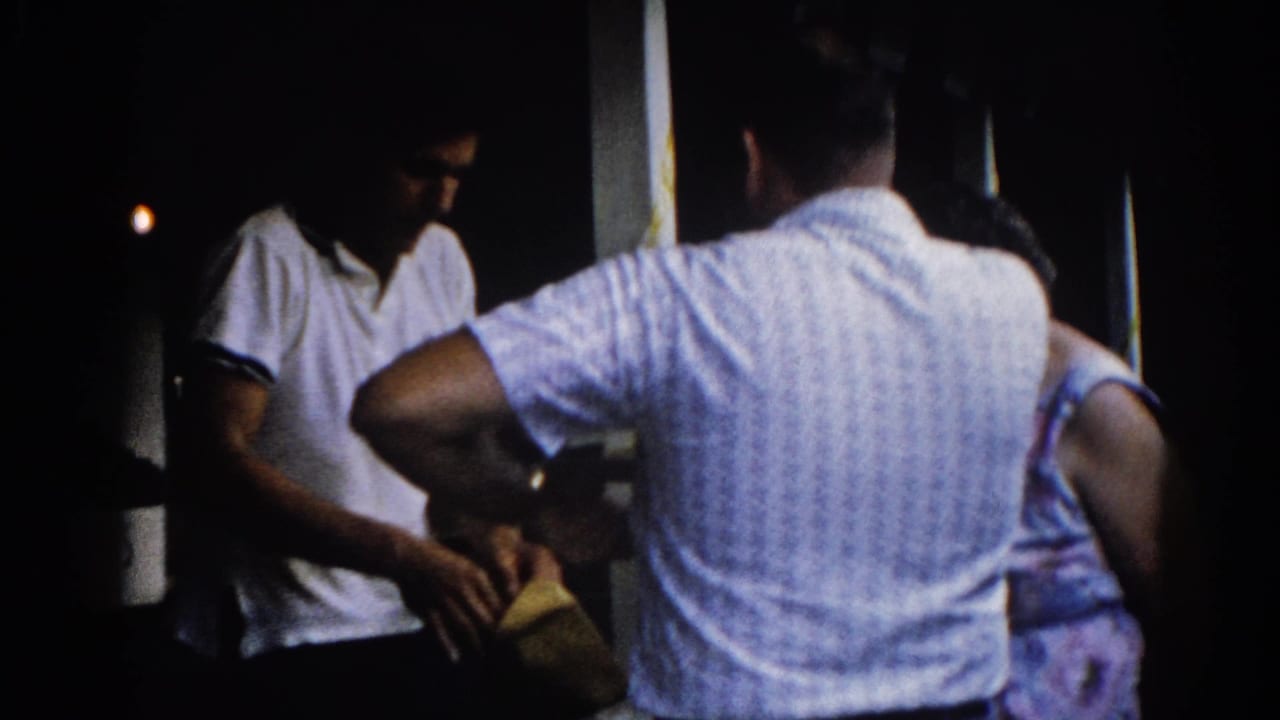 8mm home movie, Illinois circa 1962: two men in summer casualwear on a dimly lit outdoor porch at night, a yellow envelope visible in hand.