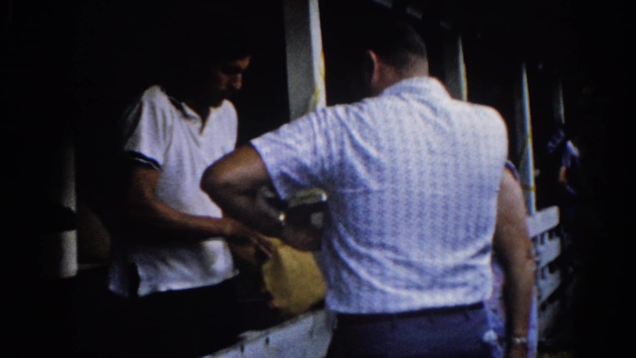 Two men on a dark porch, the nearer man's back to camera in a patterned shirt, the other in a white polo holding a yellow envelope.