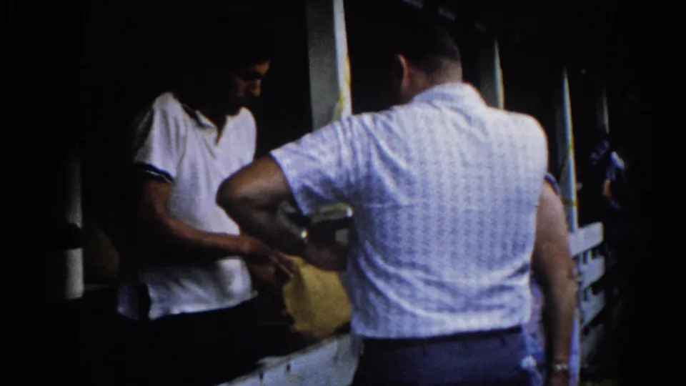 8mm home movie, Illinois circa 1962: two men on a night porch, the nearer figure turned away as the other holds a yellow envelope; partial figure visible at right.