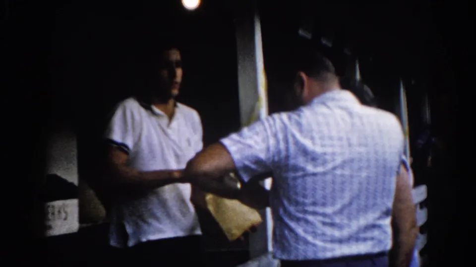 8mm home movie, Illinois circa 1962: two men conversing on a night porch, with a partial business sign reading BERKS at the left frame edge.
