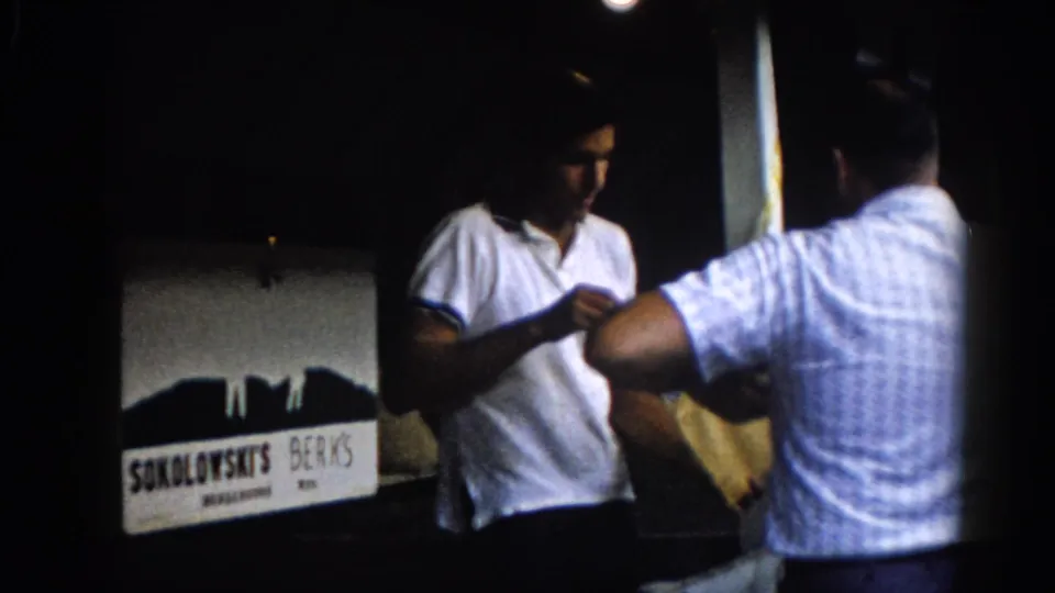 8mm home movie, Illinois circa 1962: two men exchanging a yellow envelope on a night porch, the SOKOLOWSKI'S BERKS business sign clearly visible behind them.