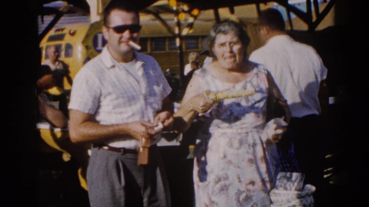 Man in white shirt and sunglasses stands beside an older woman in a floral dress at a crowded fairground, yellow vehicle behind them