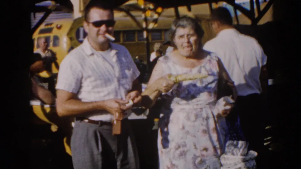 Man in white short-sleeve shirt and sunglasses stands beside an older woman in a floral dress at a 1962 Illinois fairground; yellow school bus visible in background