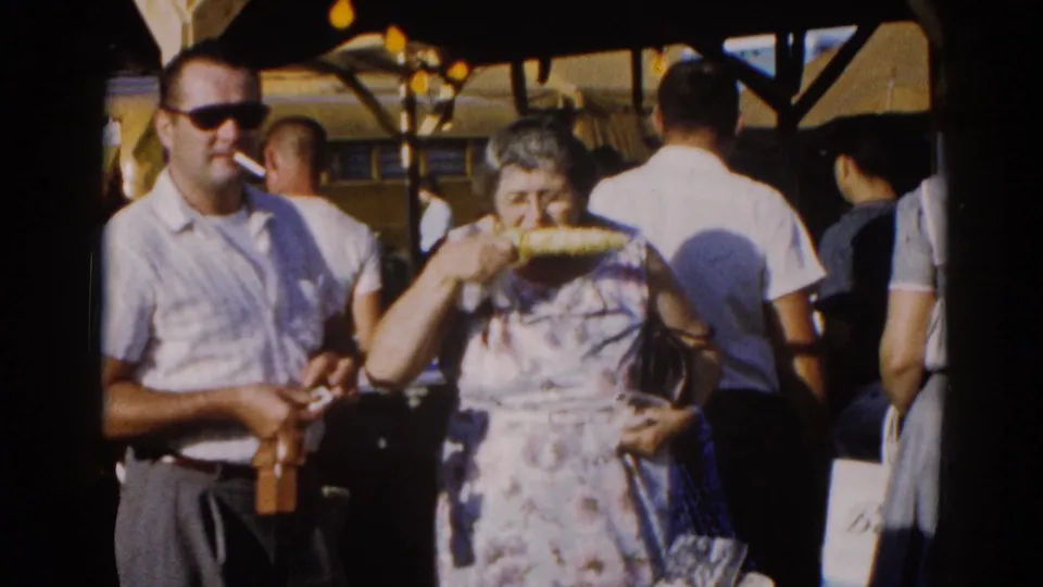 Man smoking a cigarette and woman eating corn on the cob in 1960s clothing amid a crowded Illinois fair crowd, circa 1962; yellow bus visible behind