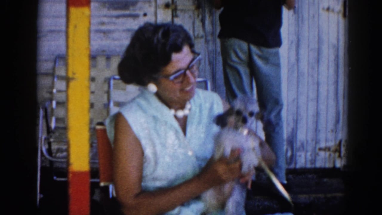 Woman in 1960s shift dress and cat-eye glasses holds small dog while a second person stands behind her against weathered wood siding, Illinois, circa 1962.