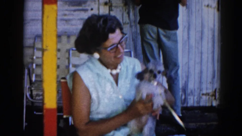 Woman in 1960s shift dress and cat-eye glasses holds small dog while a second person stands behind her against weathered wood siding, Illinois, circa 1962.