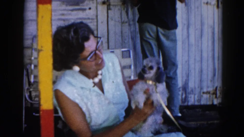 Woman with bouffant hair, cat-eye glasses and pearl necklace gazes affectionately at small dog she cradles; 8mm home movie, Illinois, circa 1962.