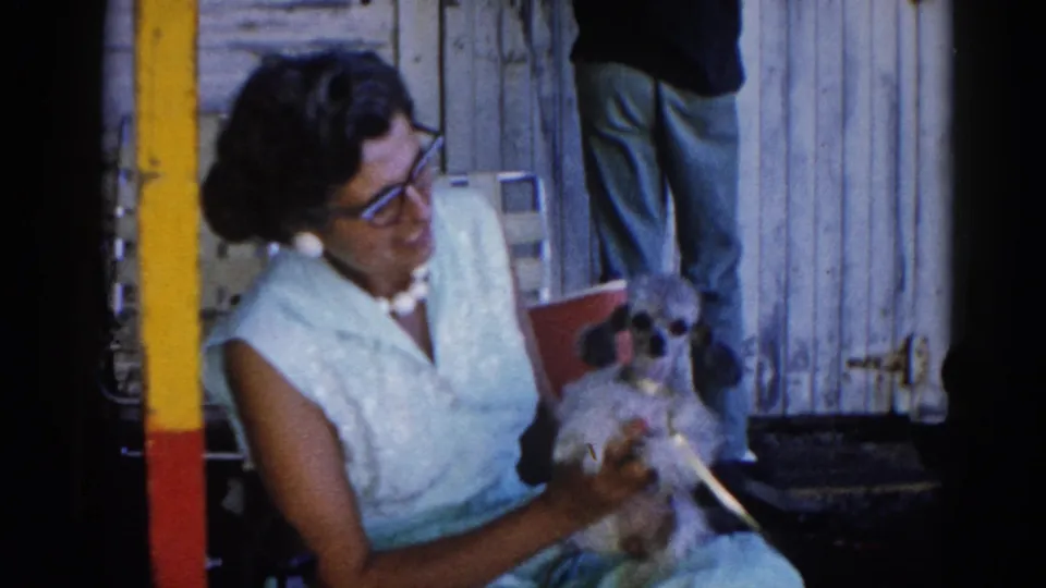 Final frame of 8mm sequence shows woman in light blue 1960s dress cradling a small dog against a weathered outbuilding wall, Illinois, circa 1962.