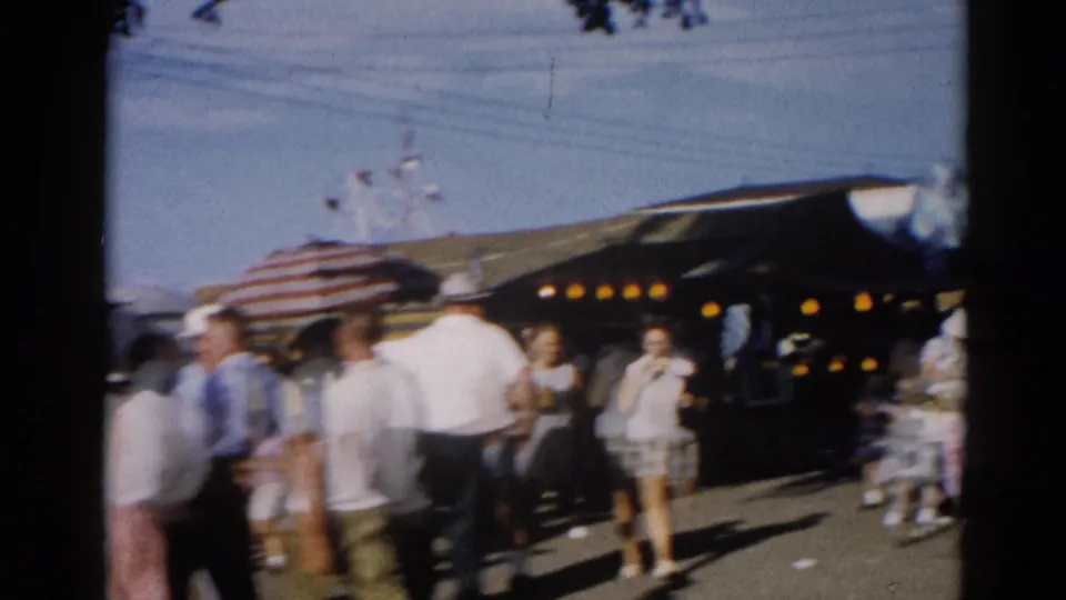 Outdoor crowd scene, possibly a county fair, with an American flag and illuminated tent structure; filmed in Illinois circa 1962 on 8mm home-movie film.