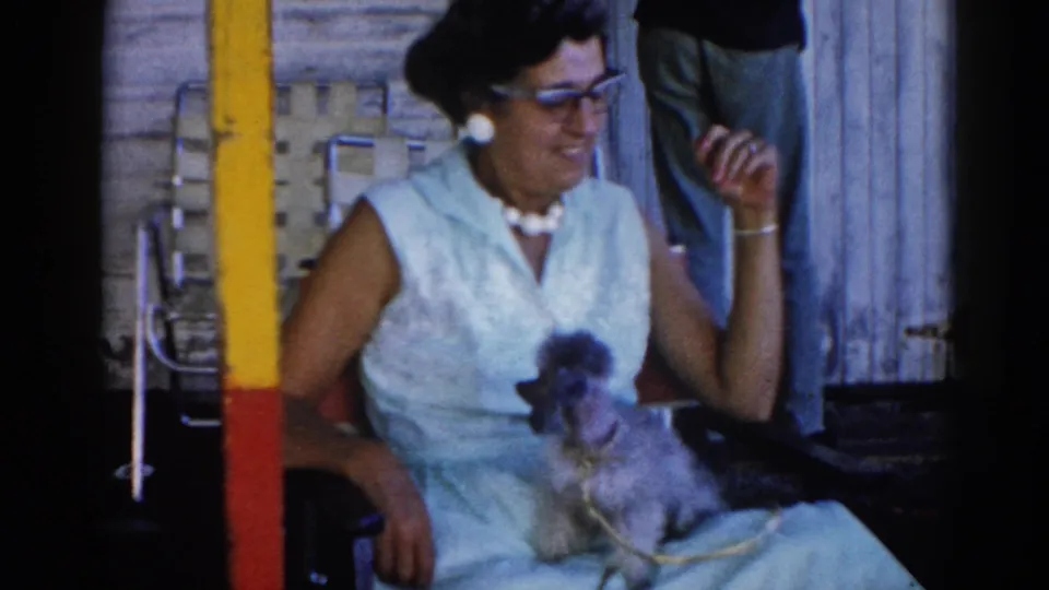 Woman in light blue 1960s sleeveless dress and cat-eye glasses smiling with gray poodle; outdoor setting, Illinois, circa 1962, 8mm home-movie film.