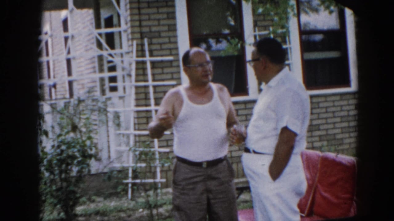 Two men in a sunlit backyard conversing; one in a white tank top and glasses, the other in a white short-sleeved shirt, with a ladder against a brick house behind them.
