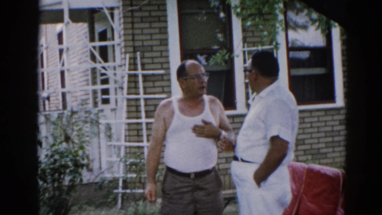Man in white tank top and glasses gestures during outdoor conversation beside a brick house; a white ladder is visible leaning against the wall.