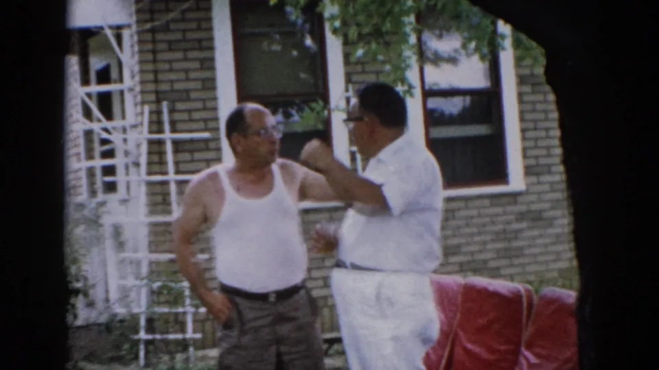Two men in animated backyard exchange, one gesturing with arm extended; brick house, white ladder, red patio furniture, and summer foliage visible, Illinois circa 1962.