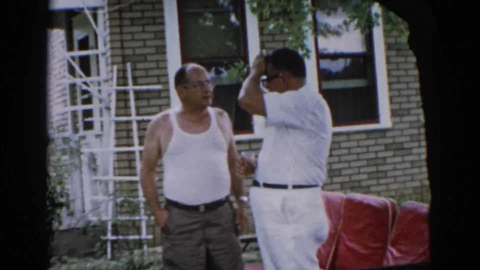 Man in white tank top pauses in backyard conversation outside a brick home in Illinois, circa 1962; white ladder leans against the brick wall behind him.
