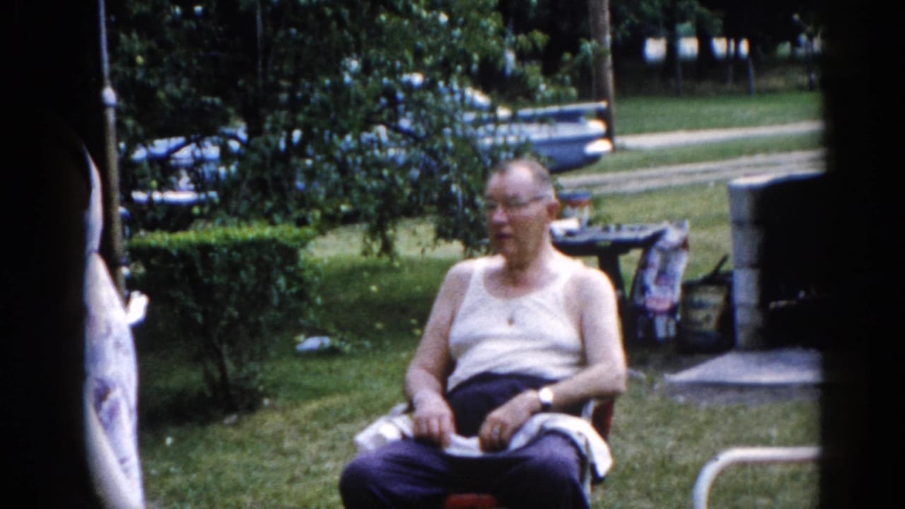Older man in white tank top and glasses seated in a folding lawn chair on a green backyard lawn, looking to one side.