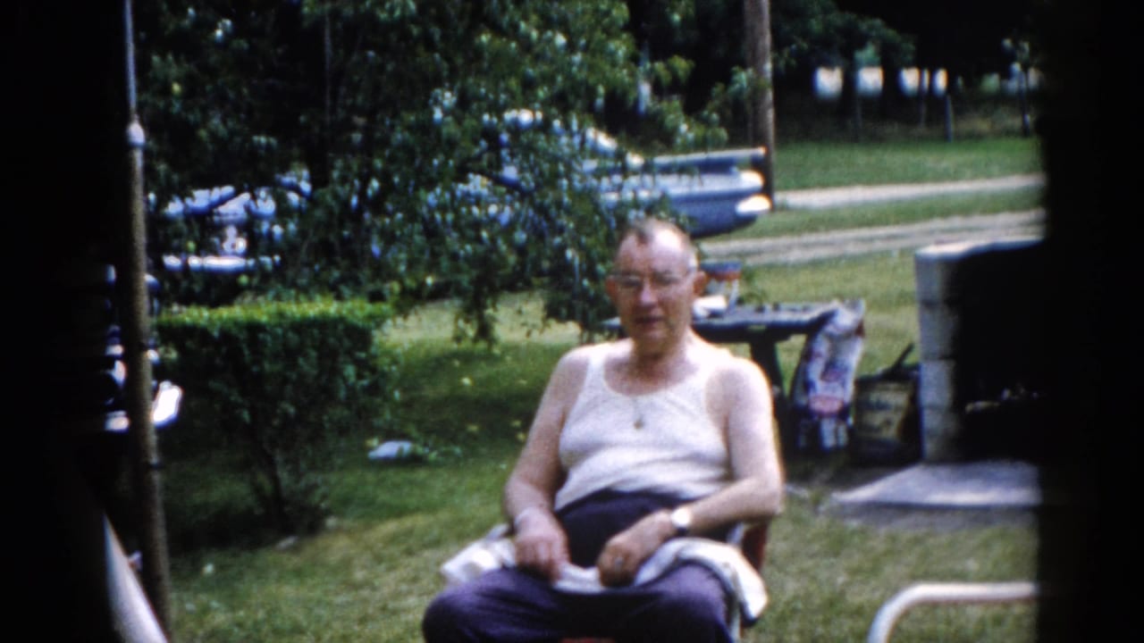 Older man in white tank top and glasses seated in a lawn chair on a green suburban lawn, turning to face the camera.