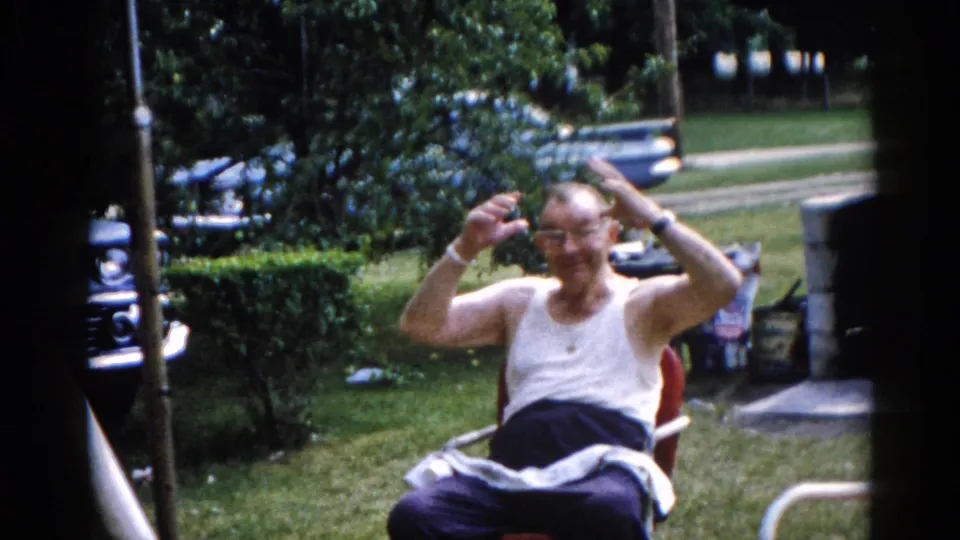 Older man in white tank top and glasses making an exaggerated animated gesture with arms raised while seated in a lawn chair outdoors; early 1960s Illinois home movie.