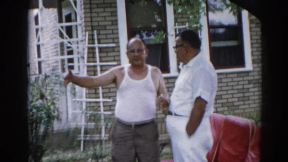 Two men near a brick building exterior — older man in white tank top with arm extended, second man in white button-up shirt; early 1960s Illinois home movie.