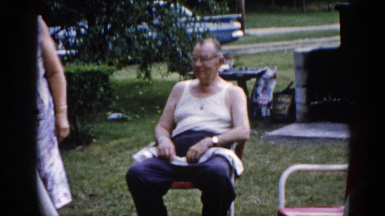 Elderly man in white tank top seated in red folding lawn chair, woman partially visible at left frame edge, green backyard with stone structure behind.