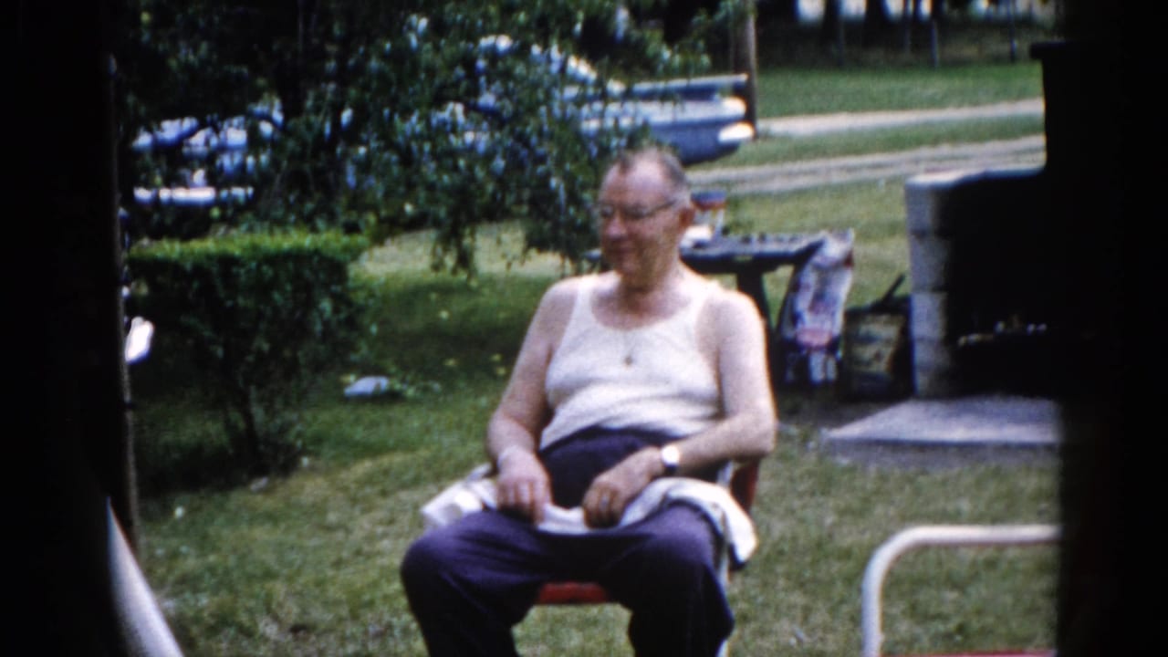 Elderly man seated in a red lawn chair in a sunny Illinois backyard, circa 1962, turning slightly and smiling; stone outdoor structure and trimmed hedges in background.