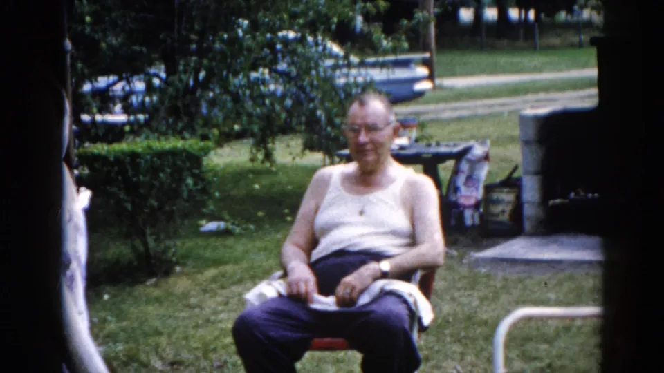Elderly man in white undershirt and glasses laughing in a red folding lawn chair, Illinois backyard circa 1962; woman partially visible at left; stone structure in background.