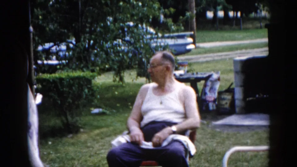 Elderly man in white undershirt, dark trousers, and glasses seated in a red folding lawn chair in a sunny Illinois backyard, circa 1962, glancing to one side.