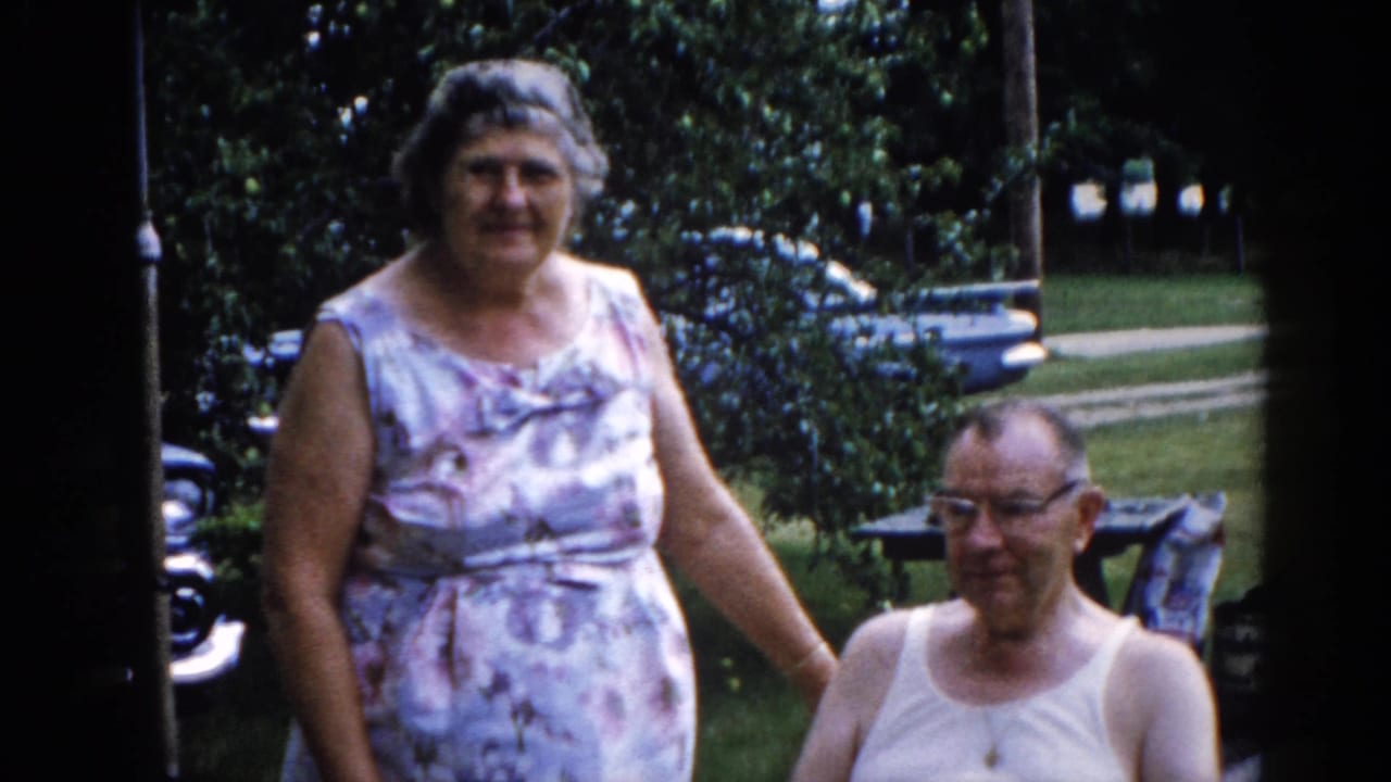 Elderly woman in floral sleeveless dress stands beside a seated man in a white tank top and glasses in a sunny backyard.