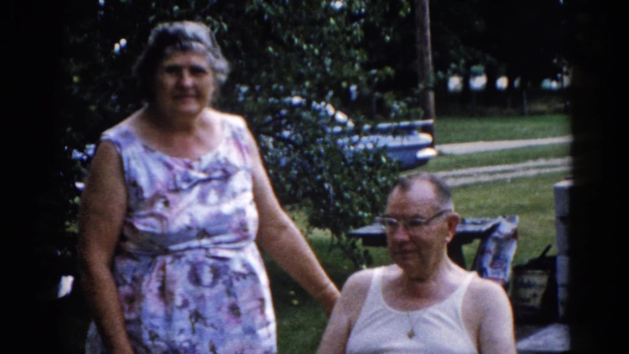 Elderly woman in patterned dress stands beside seated man with glasses in a leafy residential backyard.