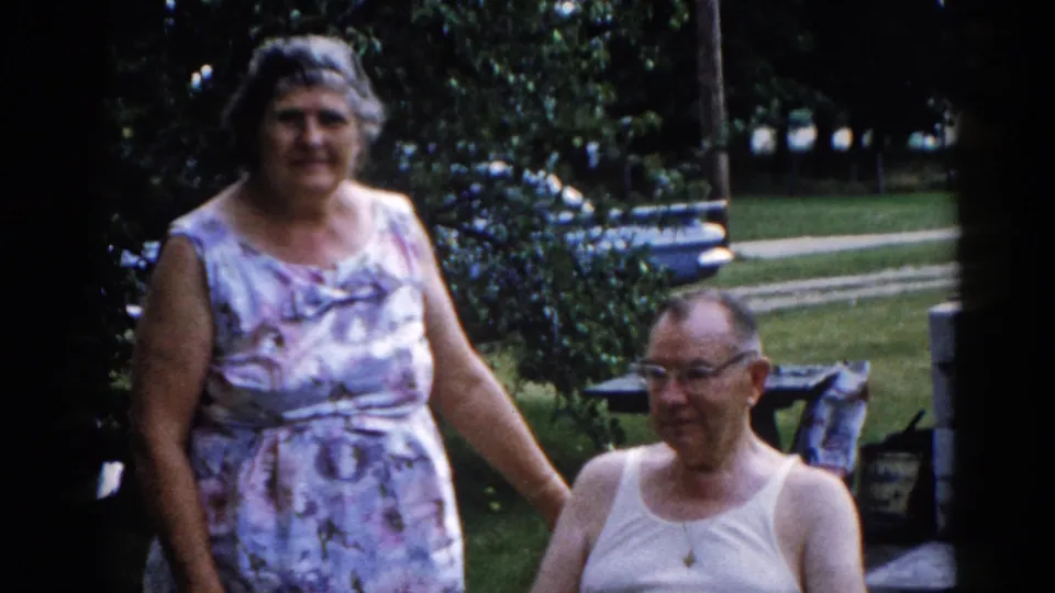 Same couple in a slightly wider frame; green lawn, leafy trees, and a background path visible, circa 1962 Illinois 8mm home movie.