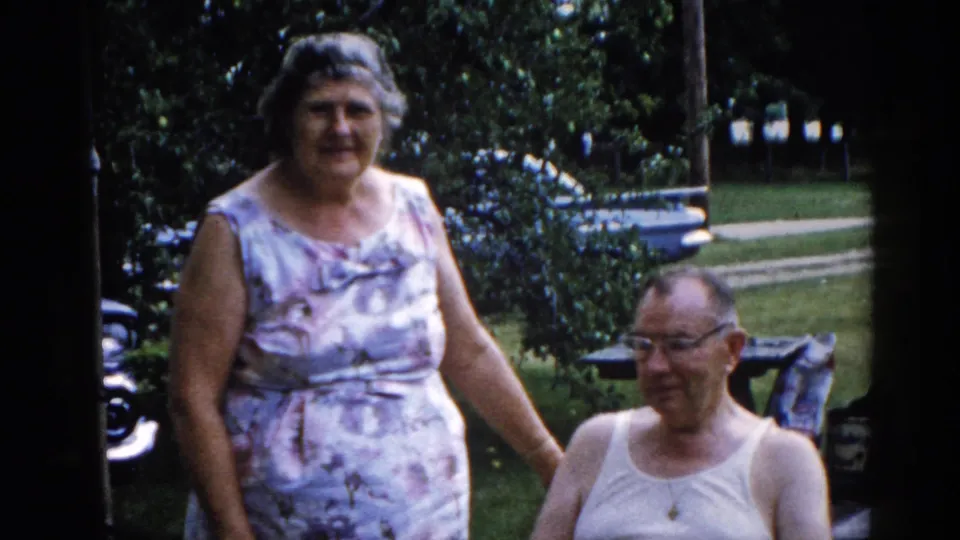Couple in a residential backyard, woman standing close to seated man; partial view of a dark car at frame left, consistent with early 1960s Illinois home movie.