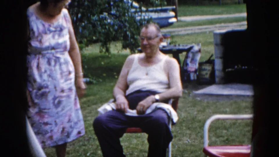 Elderly man seated in a red folding lawn chair in a sunny residential backyard, circa 1962 Illinois; wristwatch visible, woman's dress edge visible at left.