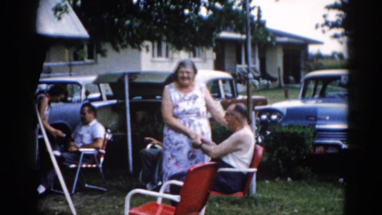 Wide backyard shot circa 1962, Illinois — elderly woman in floral dress beside a man in a white tank top in a red chair, with late-1950s American cars and a light-sided ranch house in background.