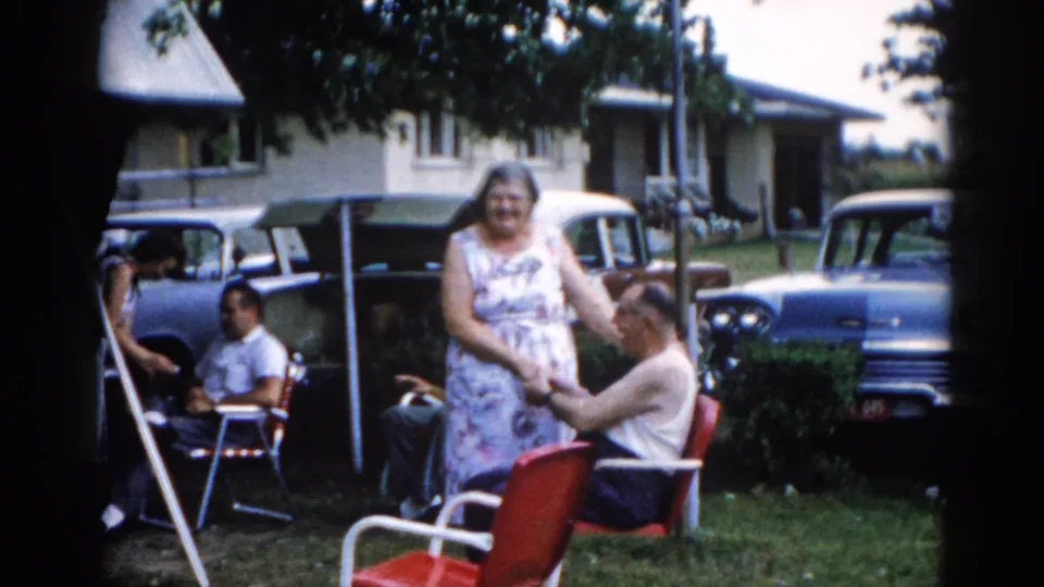Wide backyard shot circa 1962, Illinois — elderly woman in floral dress beside a man in a white tank top in a red chair, with late-1950s American cars and a light-sided ranch house in background.