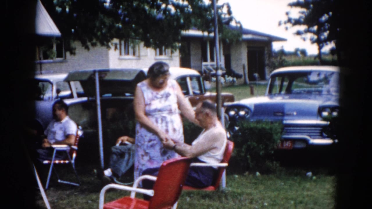 Backyard family gathering circa 1962, Illinois — elderly couple near red metal lawn chairs, with late-1950s American sedans and a light-sided ranch house behind them, younger family member at left.