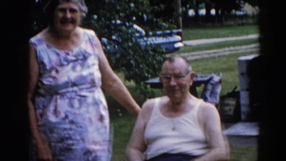 Close-up of elderly couple holding hands in a backyard, circa 1962 Illinois — woman in floral patterned sleeveless dress smiling, man wearing glasses and white undershirt seated beside her.
