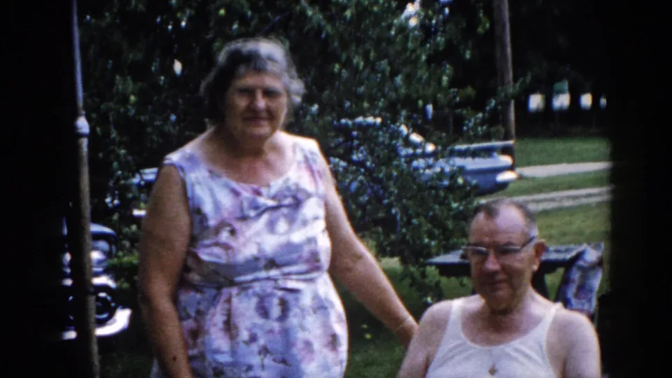 Close-up of elderly couple smiling together in a suburban backyard, circa 1962 Illinois — woman in patterned sleeveless dress, man wearing glasses and white undershirt, green shrubbery behind them.