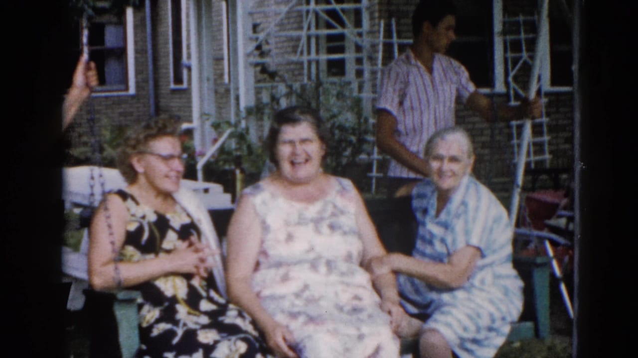 Three adult women laughing on a porch swing in a backyard garden, Illinois, 1962; a man in a striped shirt stands in the background near the trellis.