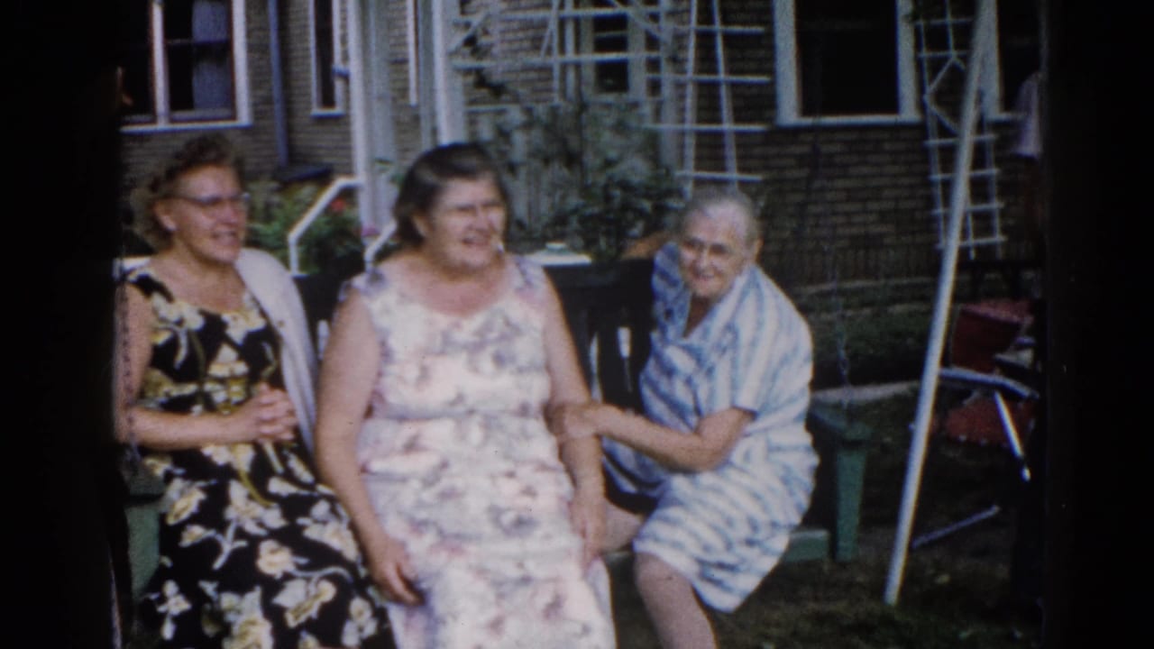 Two smiling women in floral dresses on an outdoor porch swing, with a third older woman at right in a light blue garment; suburban backyard, Illinois circa 1962.