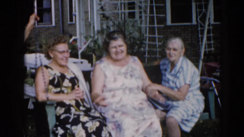 Three adult women in 1960s-era summer dresses seated on a porch swing in a suburban backyard, Illinois, approximately 1962.